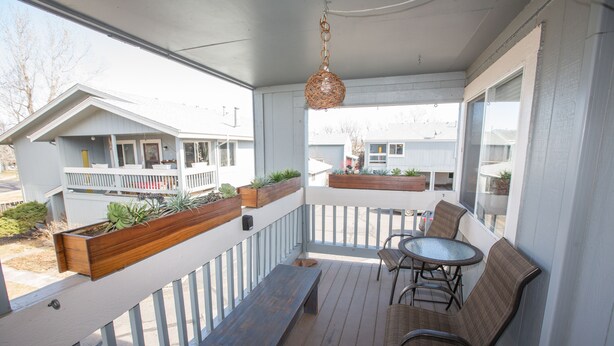 Une terrasse extérieure couverte, aménagée avec un banc, des chaises et une table. Des jardinières avec des plantes sont disposées sur la balustrade.