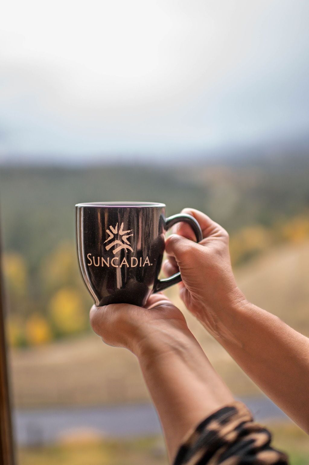 A person holding a black mug with the text "SUNCADIA" and a sun design, with a blurred outdoor background.