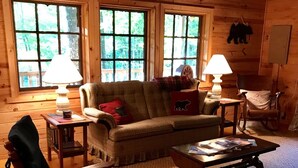 A cozy cabin interior with a rocking chair, a sofa, and a coffee table. There are two windows with a view of greenery outside. - Quiet Rustic Getaway (Whittier)