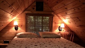 A cozy attic bedroom with a wooden ceiling, a bed with floral bedding, two wall-mounted lamps, and a window with blinds. - Quiet Rustic Getaway (Whittier)