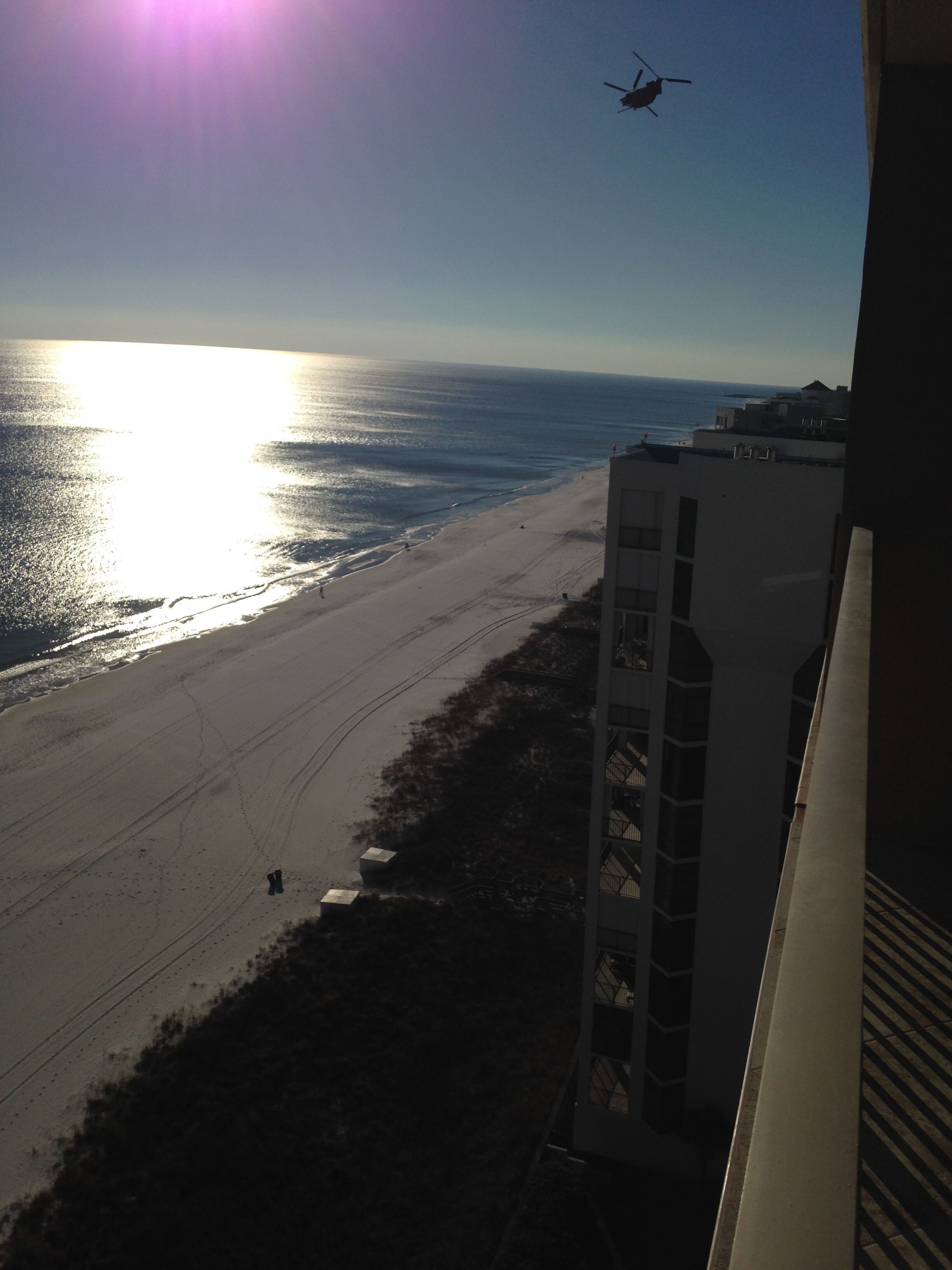 Vista de la playa con un helicóptero en el cielo, edificios en primer plano y el océano al fondo.