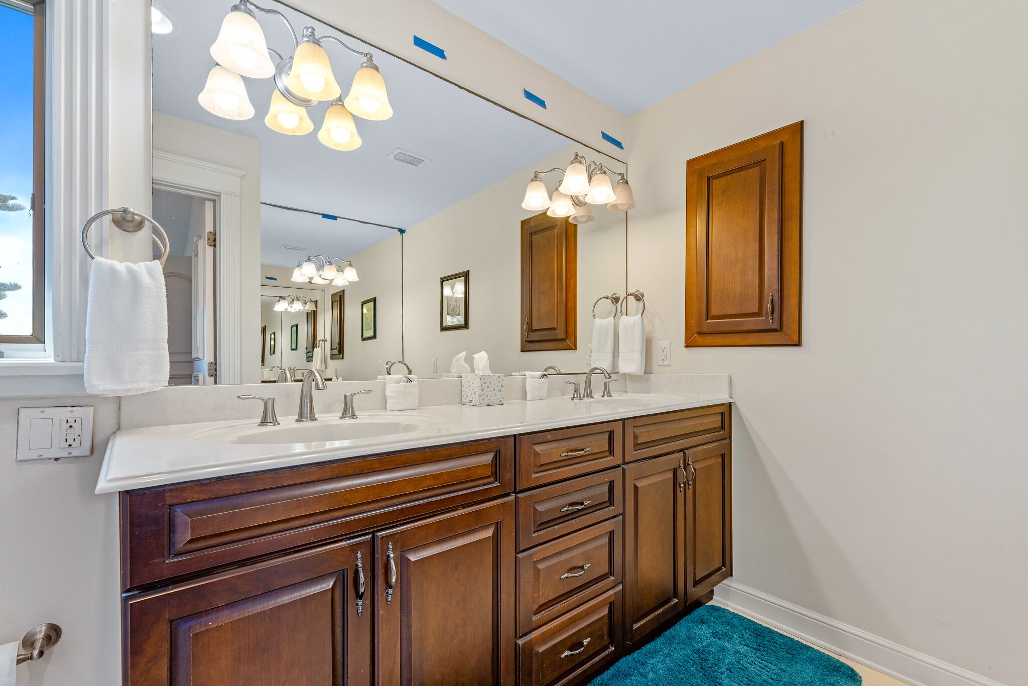 A bathroom with a large mirror, two sinks, wooden cabinets, and a blue rug.