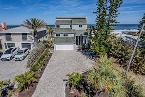 A two-story house with a driveway leading to a garage, surrounded by various plants and a view of the ocean.