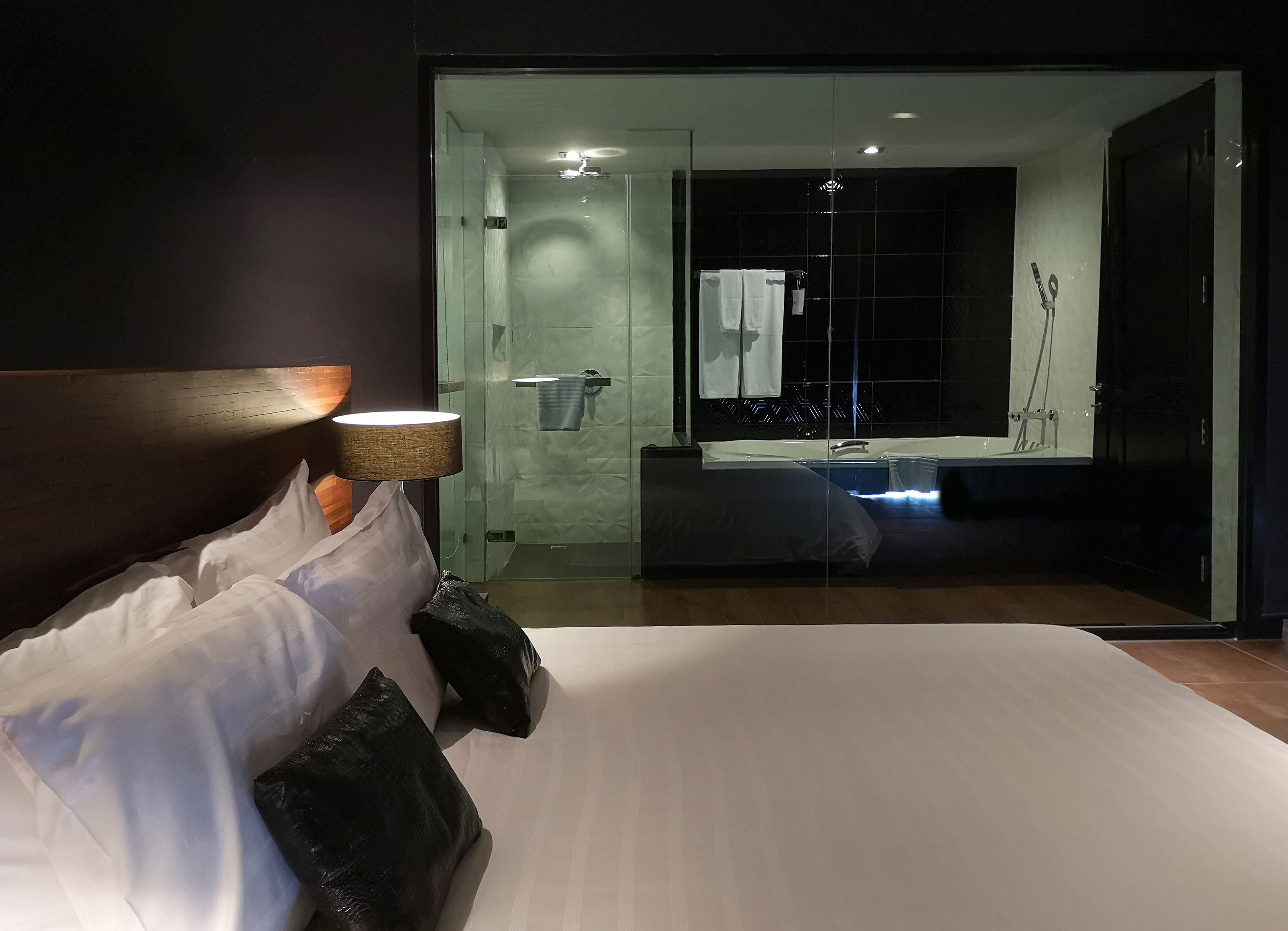 Une chambre d’hôtel moderne dotée d’un grand lit, d’une table de chevet avec une lampe, d’une douche en verre et d’une salle de bain avec une baignoire et un miroir.