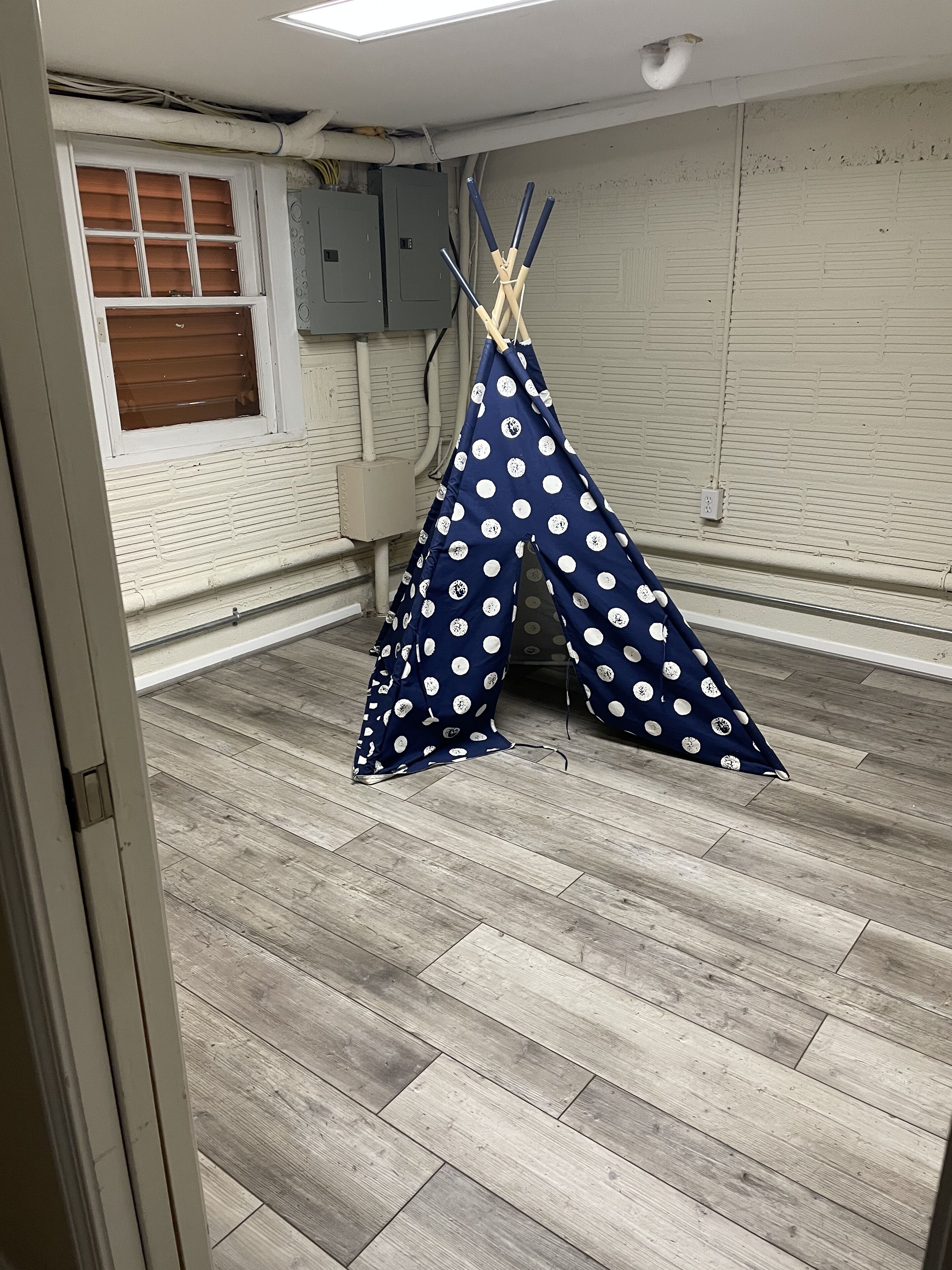 A room with a wooden floor, a window with blinds, and a blue and white polka dot teepee tent.