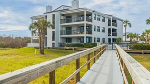 A modern multi-story building with balconies, a wooden boardwalk leading to the entrance, and a grassy area with palm trees. - Bright and Spacious 3 bedroom ocean front, ground floor condo (St. Augustine Beach)