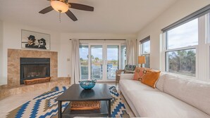 A living room with a fireplace, a ceiling fan, a sofa, a coffee table, and large windows offering a view of the outdoors. - Bright and Spacious 3 bedroom ocean front, ground floor condo (St. Augustine Beach)