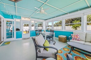 A bright, open-plan living area with a ceiling fan, a dining table with chairs, a sofa, and a window with a view of greenery.