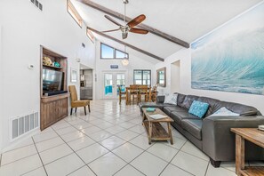 A spacious living area with a ceiling fan, a dining table with chairs, a sofa with cushions, and a large painting of a wave.