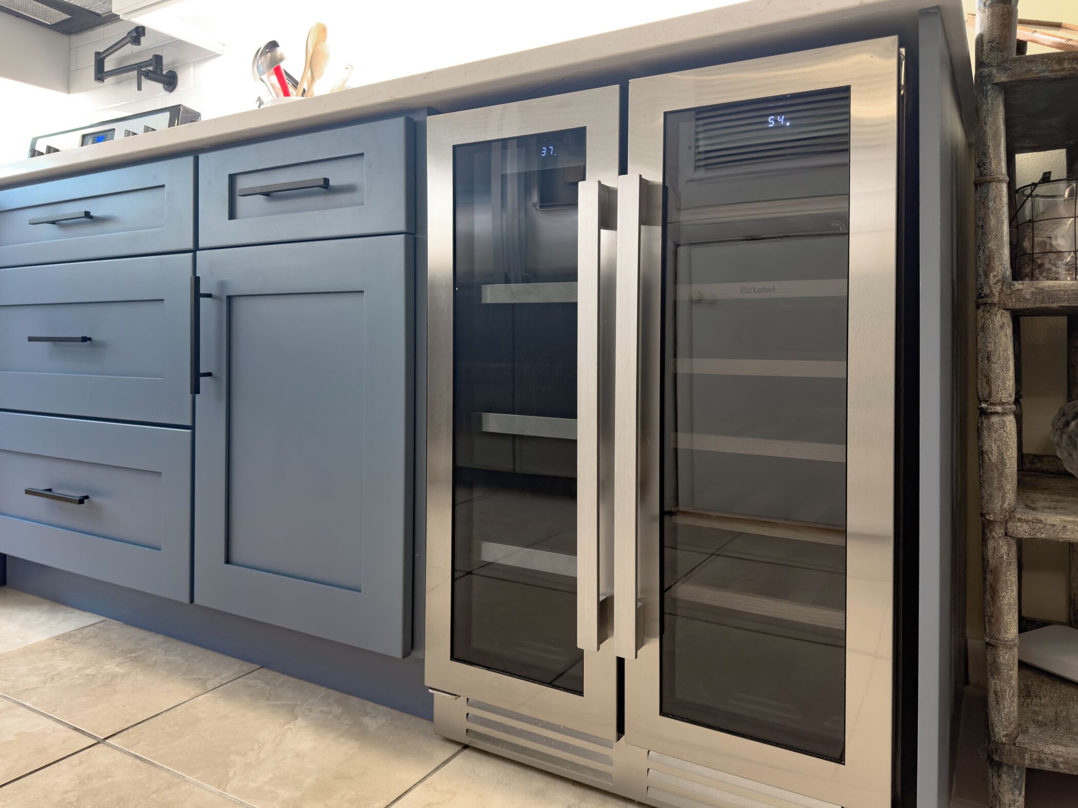 A kitchen with grey cabinets, a stainless steel refrigerator, and a wooden shelf.