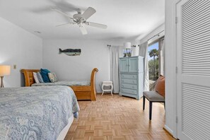 Un dormitorio con dos camas, un ventilador de techo, cabecera de madera, un taburete blanco pequeño y un armario azul claro, con vistas al exterior.