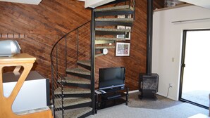 Una escalera en espiral con un televisor de pantalla plana sobre un soporte, un estufa de leña y una puerta que da al exterior.