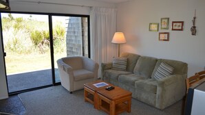 Una sala de estar con un sofá, un sillón y una pequeña mesa de madera. Hay una lámpara sobre la mesa y una ventana con cortinas.