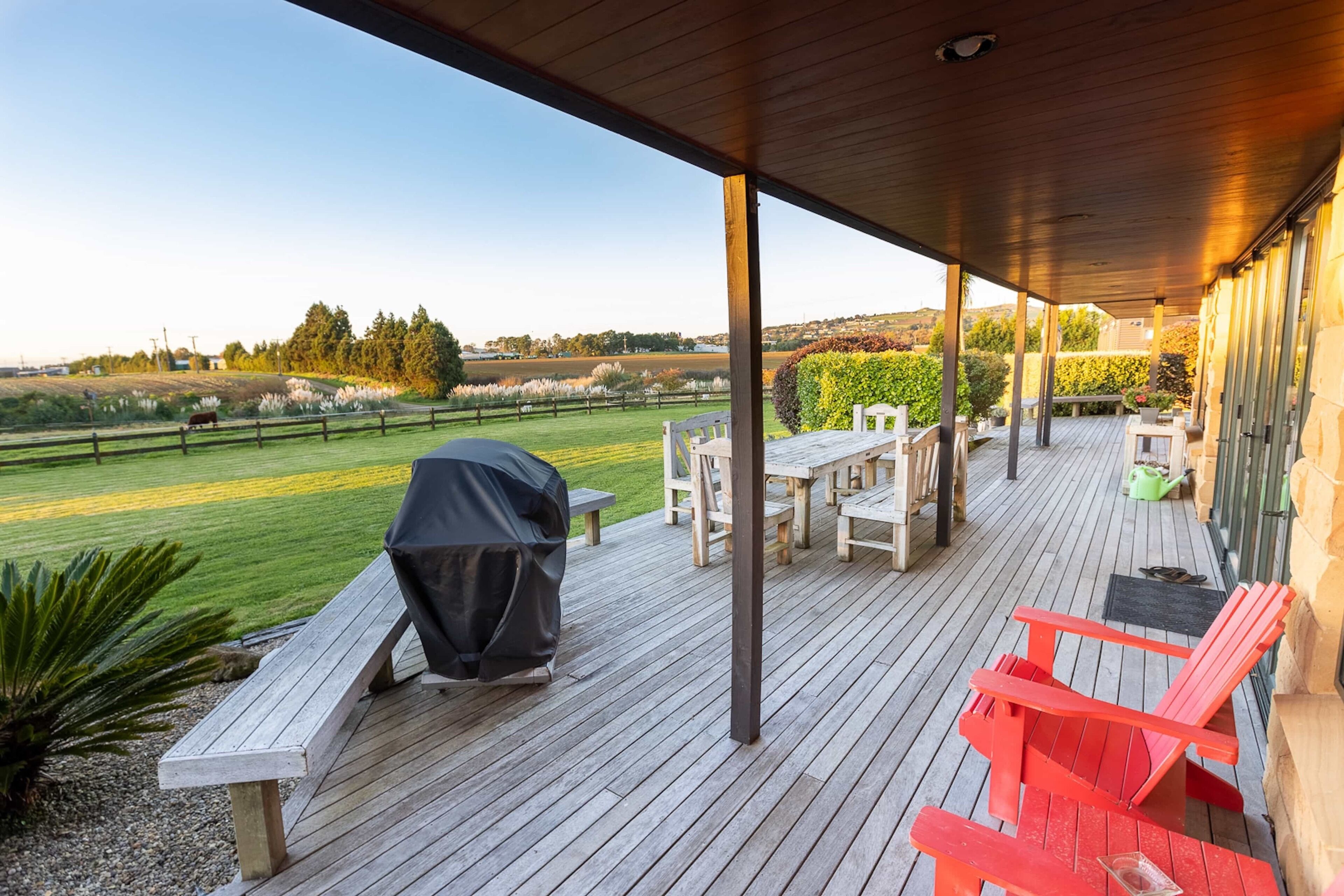 A covered outdoor deck with wooden furniture, a grill, and a view of a grassy field and trees.