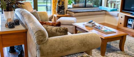 A cozy living room with a sofa, coffee table, and a TV. There is a window with a view of greenery outside.