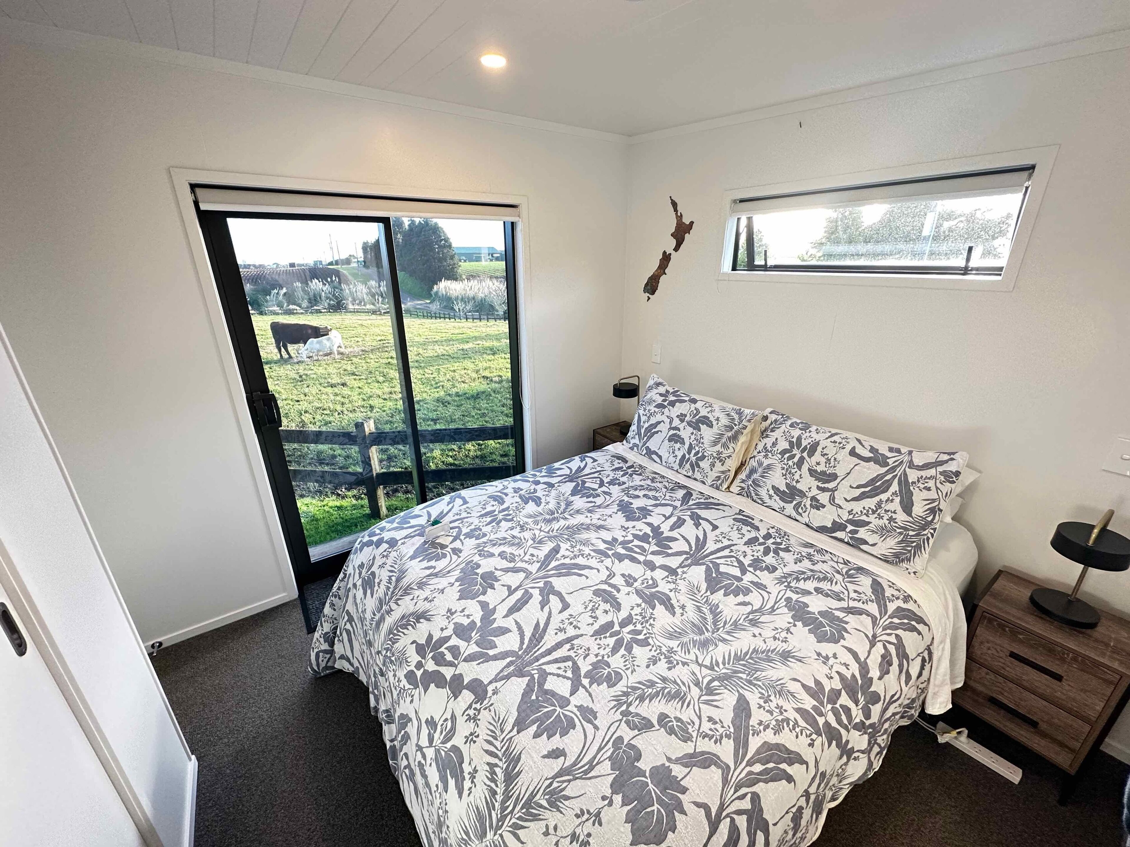 A bedroom with a large bed, a window with a view of a grassy field, a small bedside table, and a nightstand.