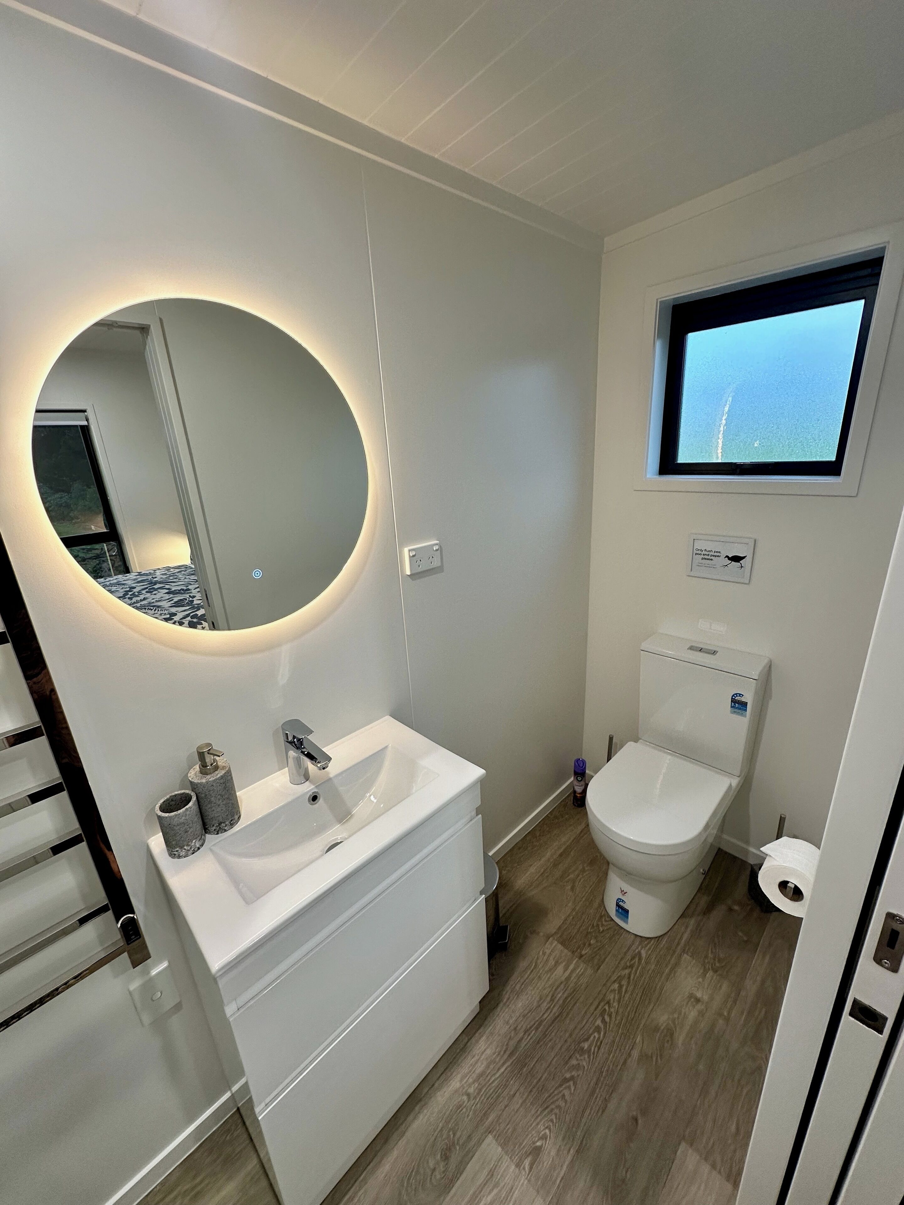 Un baño moderno con un espejo redondo, un lavamanos blanco, un inodoro y una ventana pequeña.