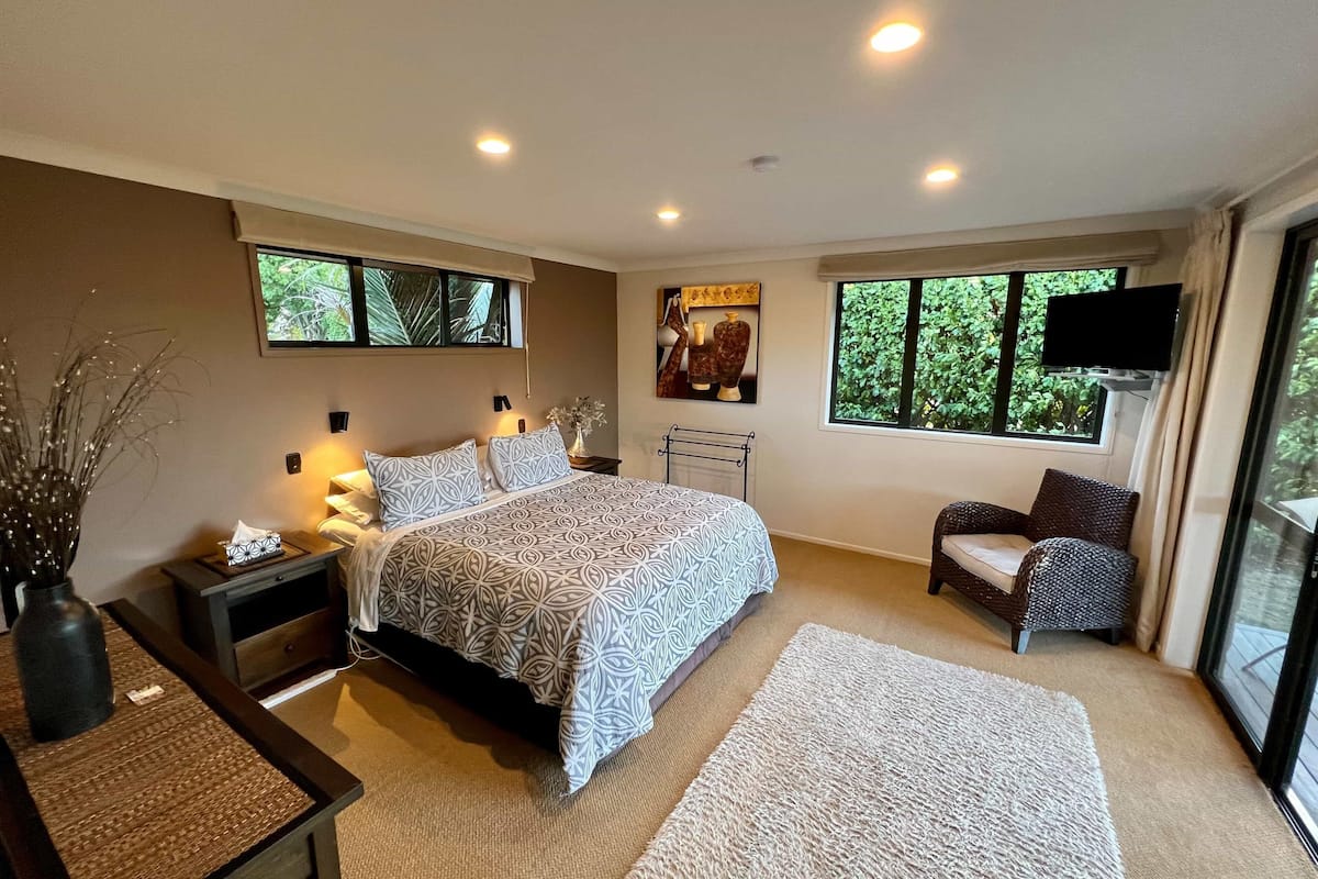 A modern bedroom with a large bed, bedside tables, a TV, and a window with a view of greenery.