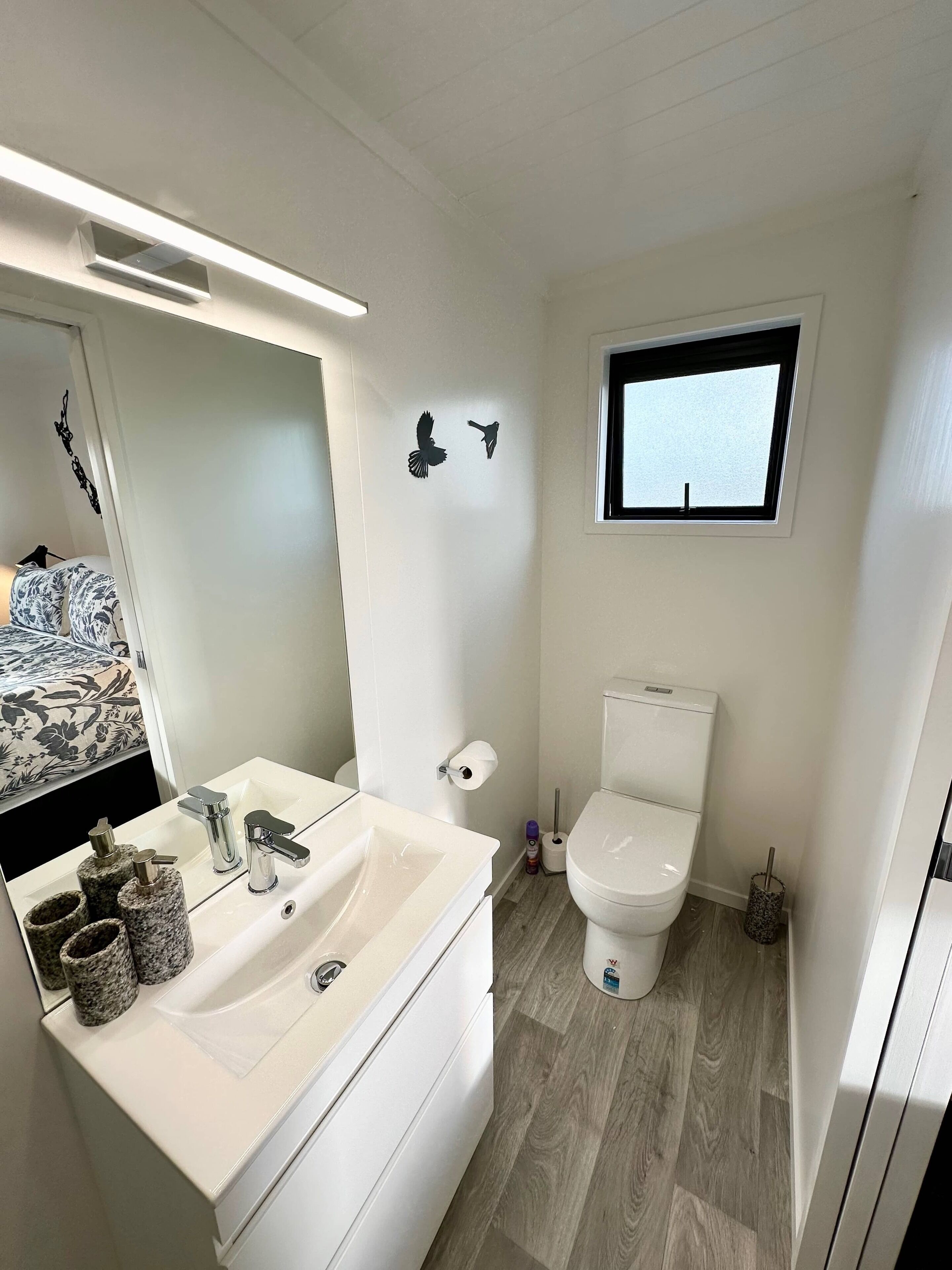 A modern bathroom with a white sink, two faucets, a mirror, a toilet, and a window.