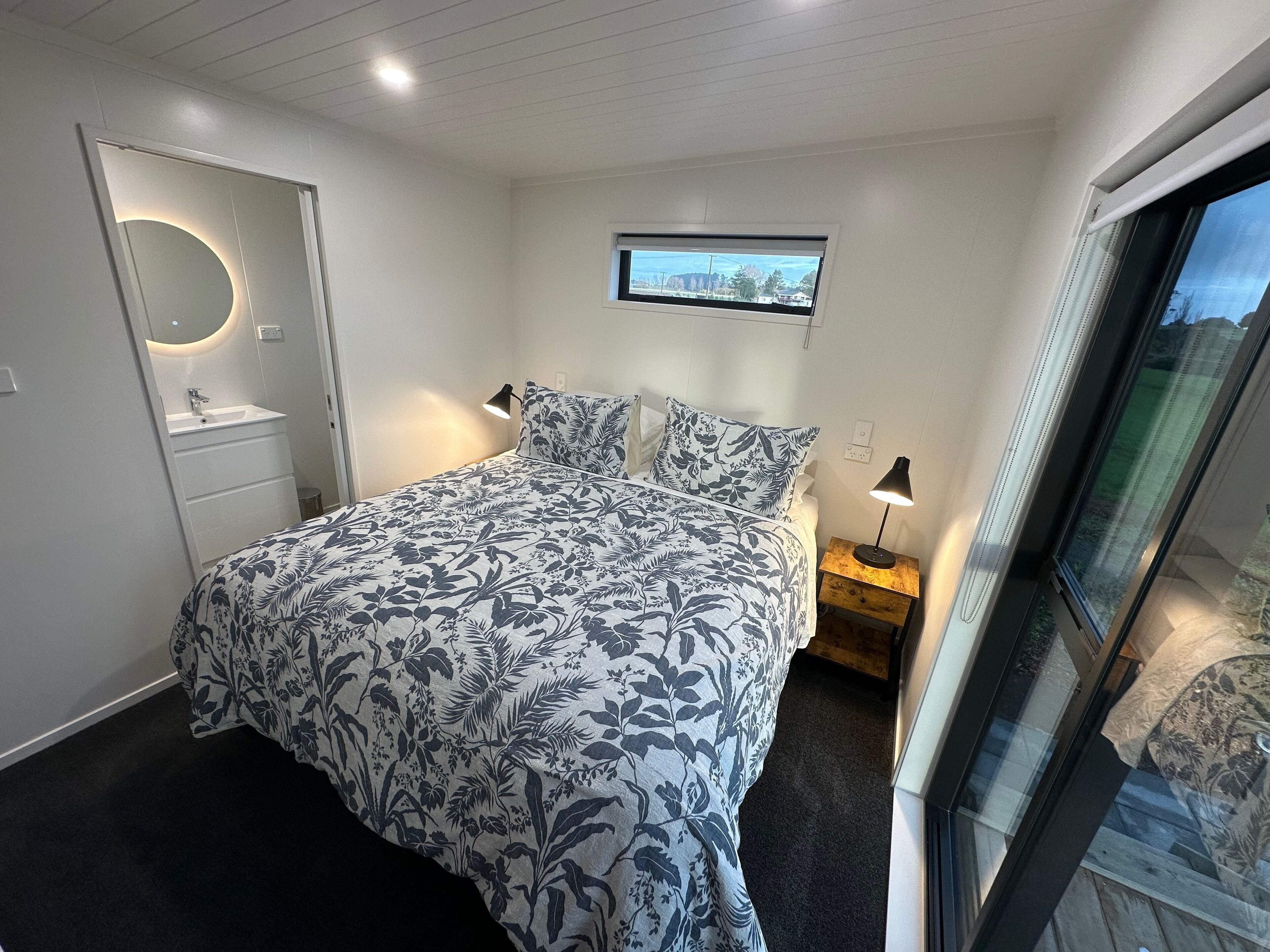A bedroom with a bed, a nightstand, a window, and a bathroom with a sink and mirror.