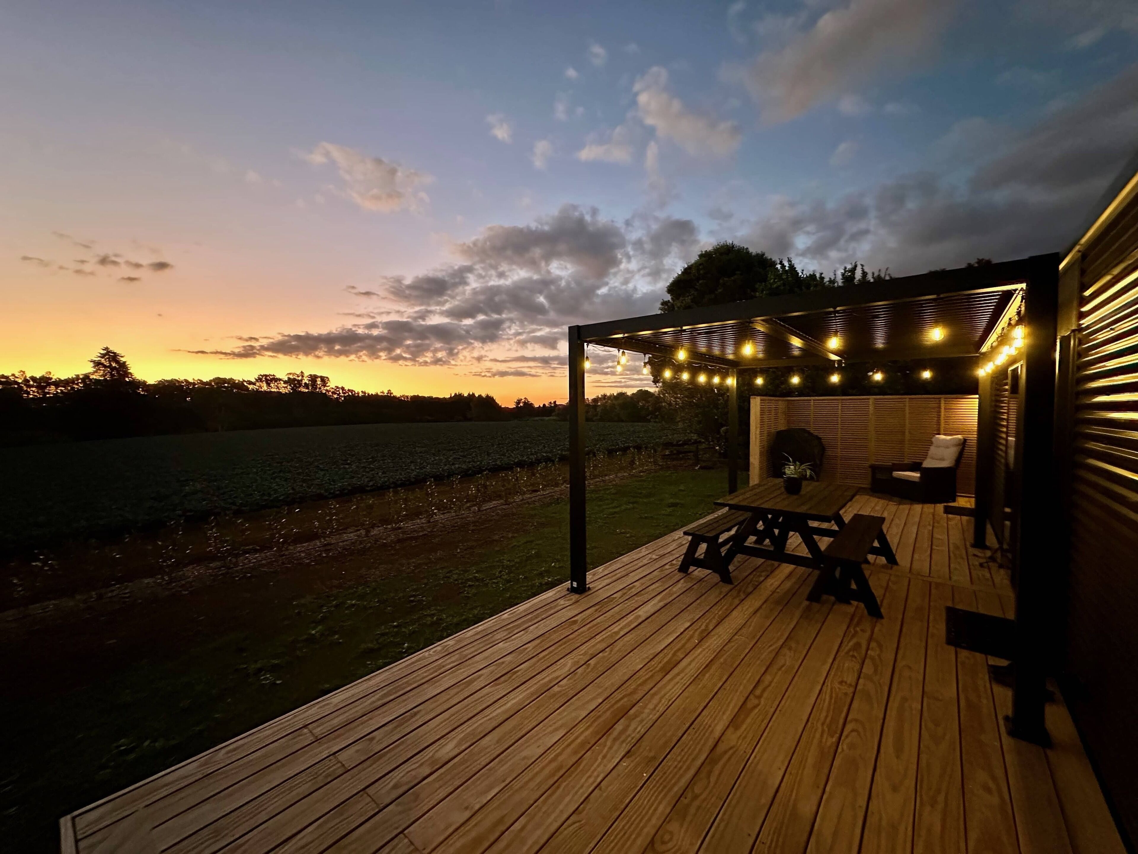 En træterrasse med pergola og lyskæder, med udsigt over et marklandskab og et vandareal.
