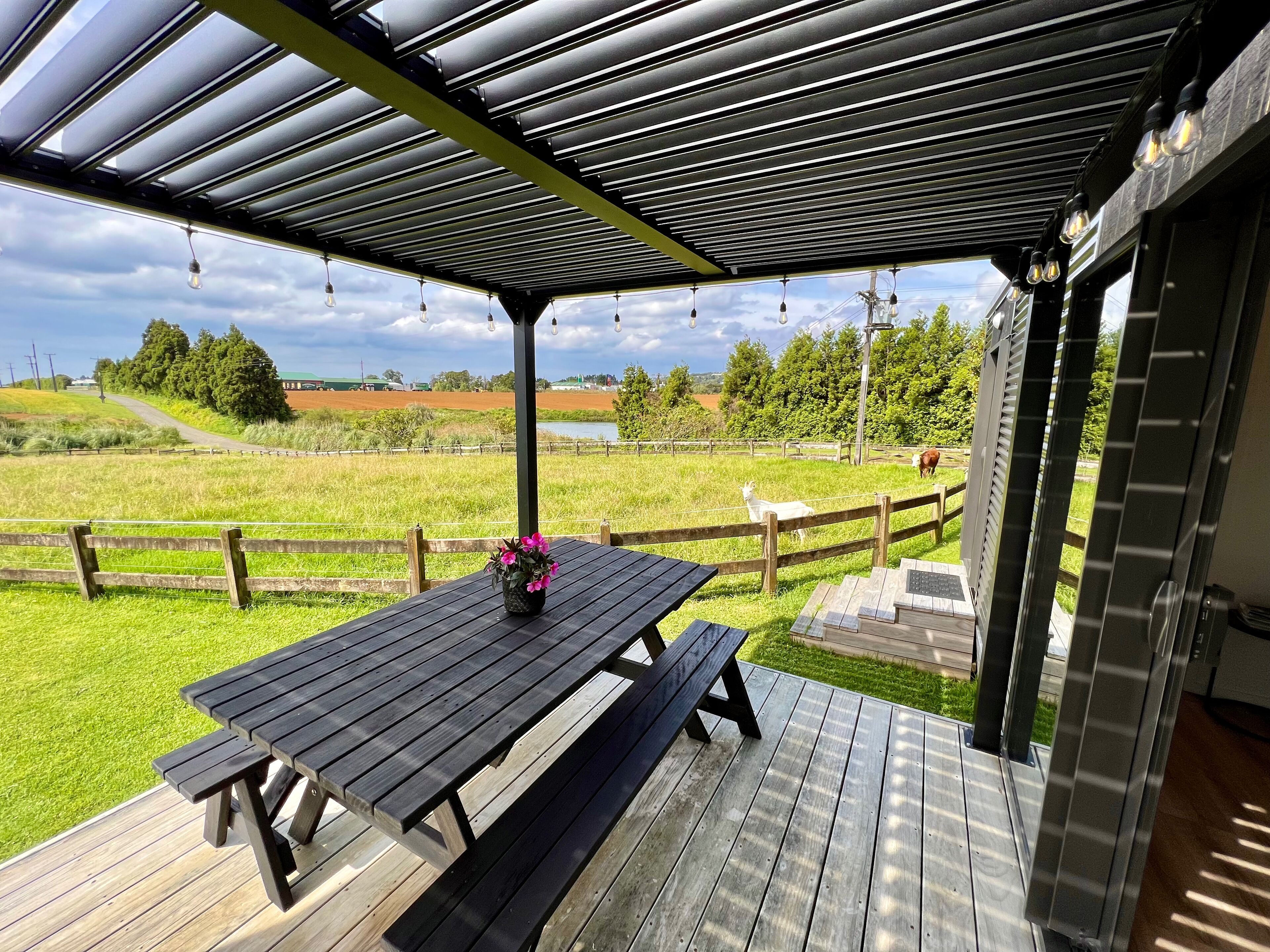 An outdoor patio with a wooden table and benches, a pergola overhead, and a view of a grassy field and trees.
