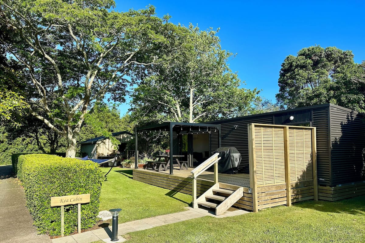 A compact, modern cabin with a covered outdoor seating area, a wooden staircase, and a neatly trimmed hedge.