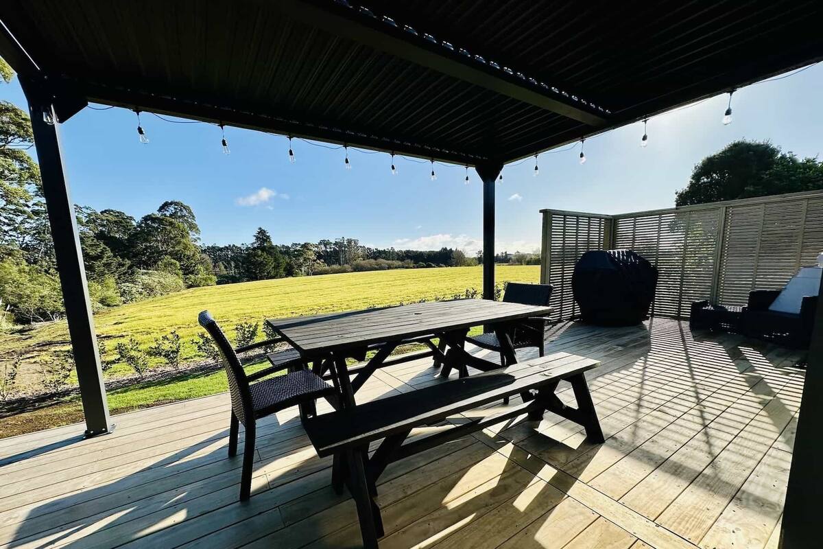 An outdoor patio with a wooden table and benches, a barbecue grill, and a view of a green field.