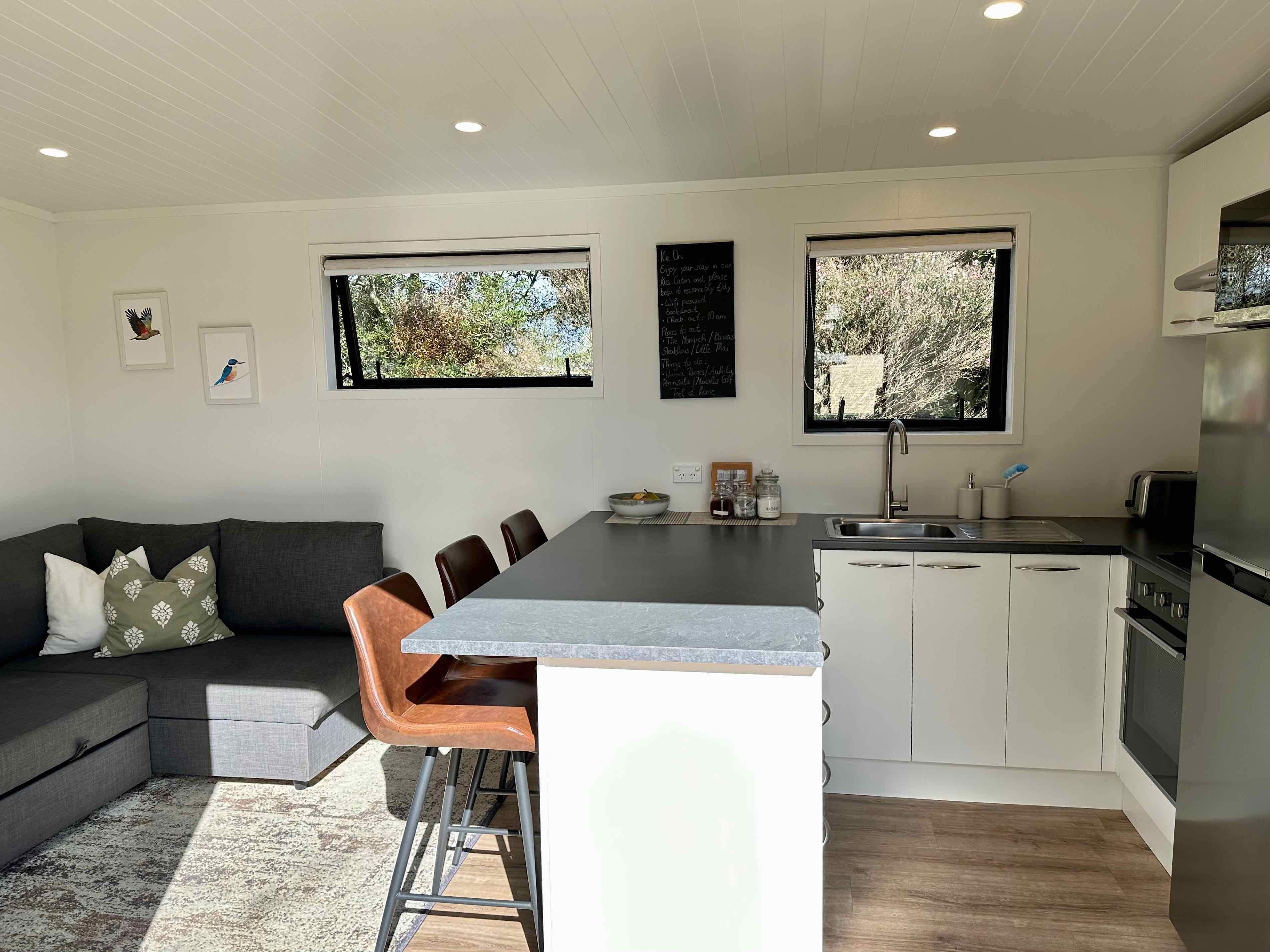 A modern kitchen with a central island, white cabinets, and a dining area with a grey sofa and brown chairs.