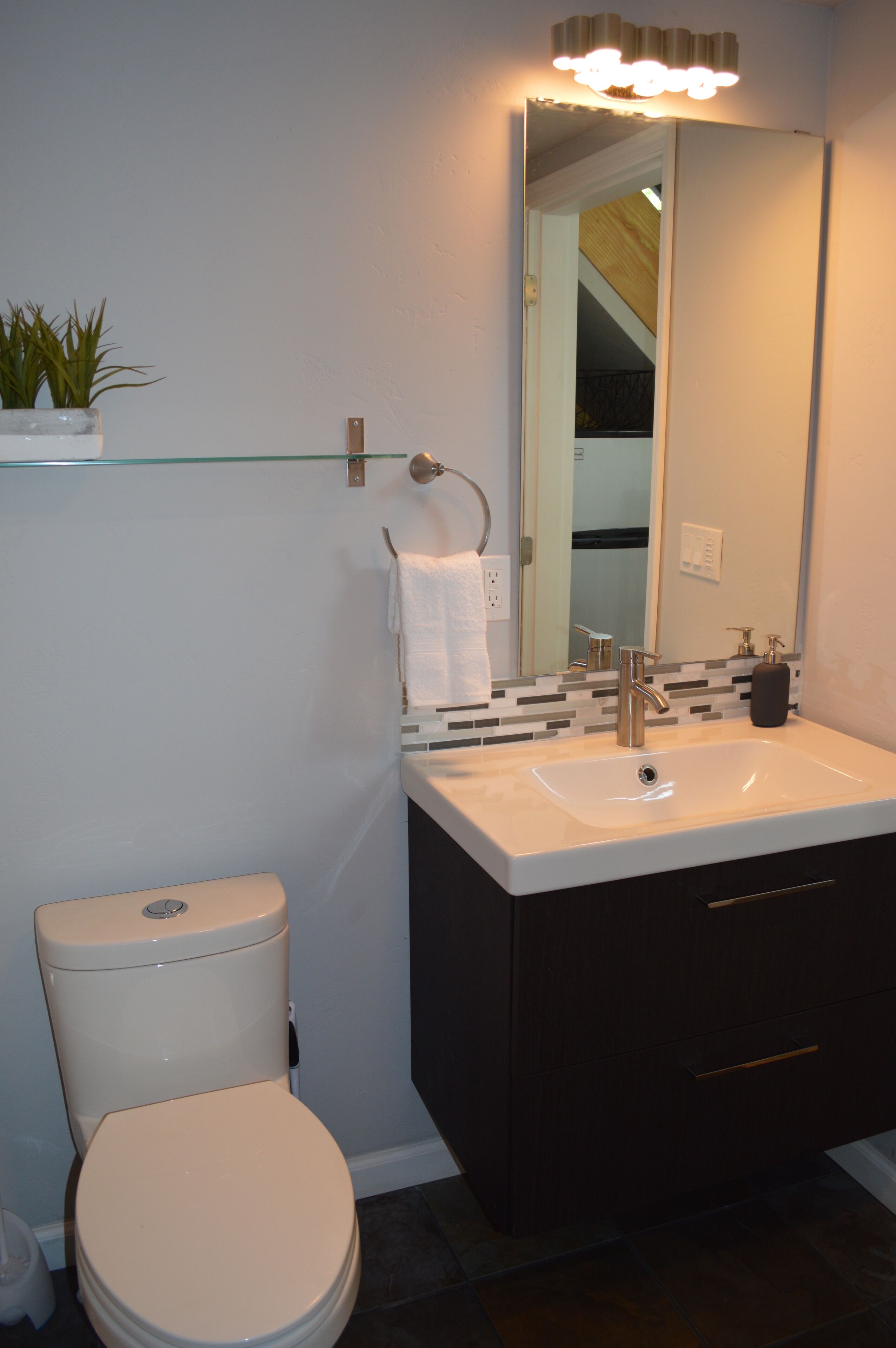 Un bagno con il water bianco, un mobile contenitore in legno scuro, uno scaffale in vetro con una pianta, uno specchio e un rubinetto moderno.