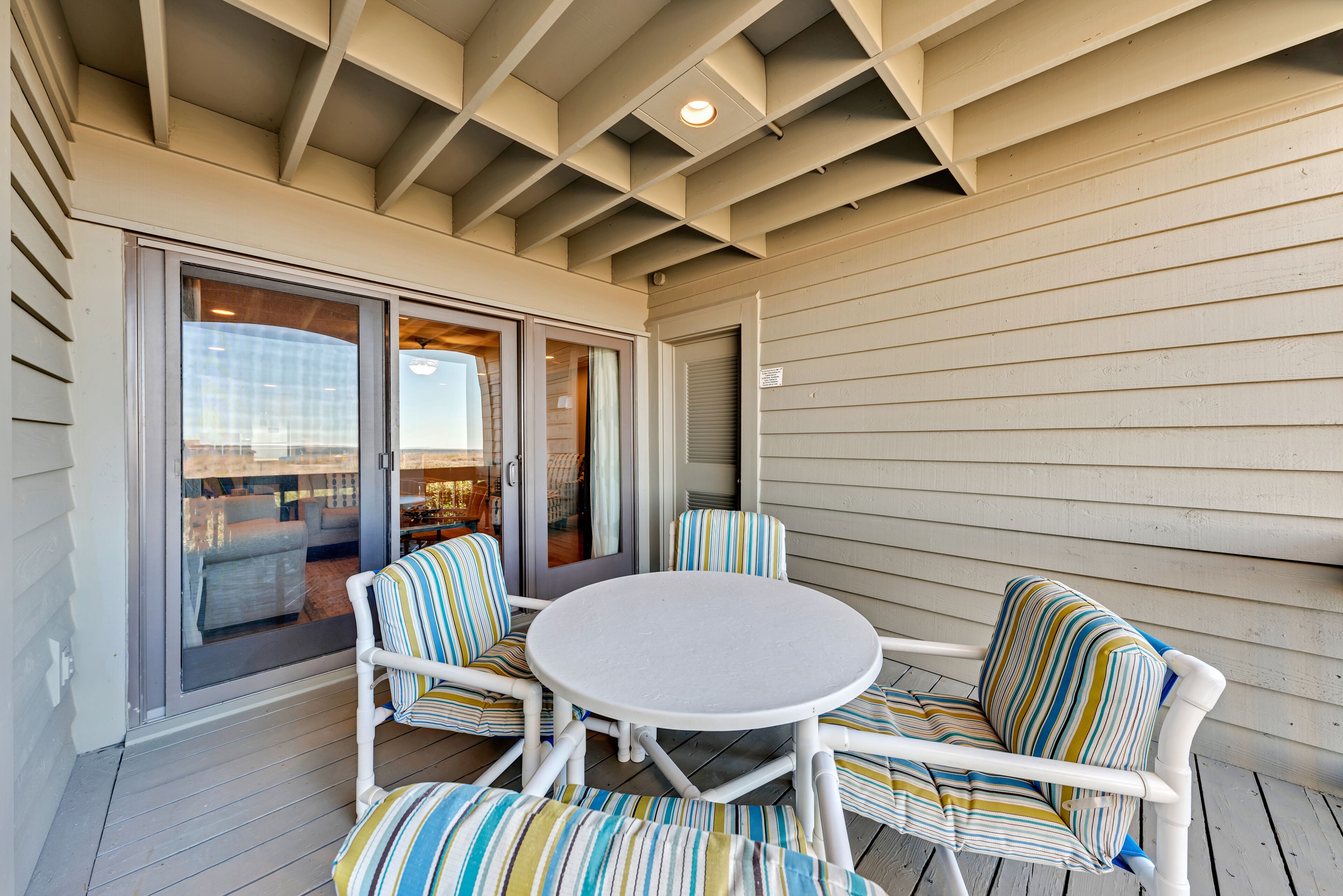 A covered patio with a round table and striped chairs, a sliding glass door leading to an interior space, and a ceiling with exposed beams.