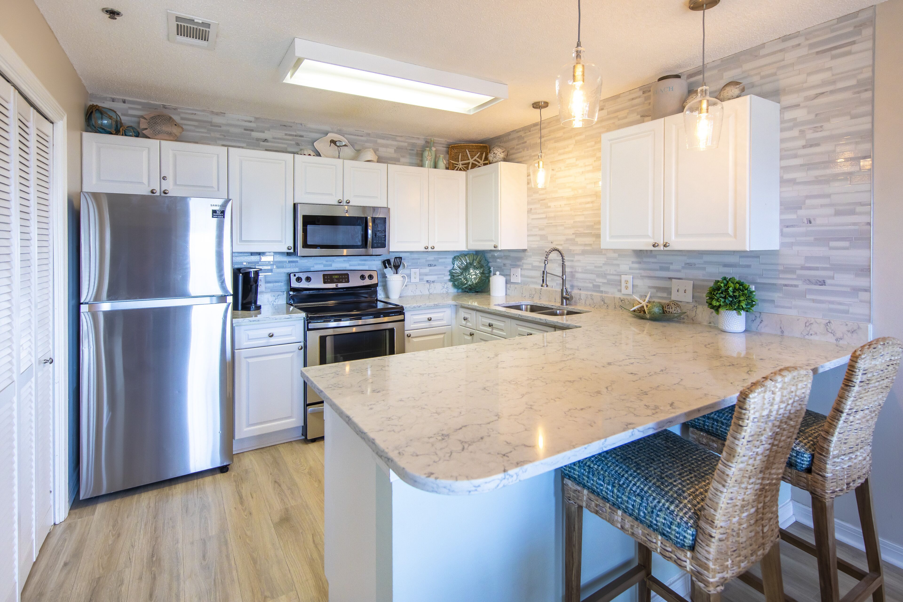 A modern kitchen with white cabinets, stainless steel appliances, a marble countertop, and wicker bar stools.