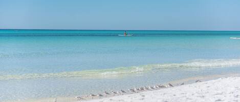 A beach with clear turquoise water, a person paddling a kayak, and seagulls on the sand.