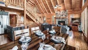 A wooden cabin interior with a dining area set for four, a stone fireplace, and a staircase leading to an upper level. - Best Panoramic Views! Hot Tub | Fire Pit | Top 10% (Sevierville)