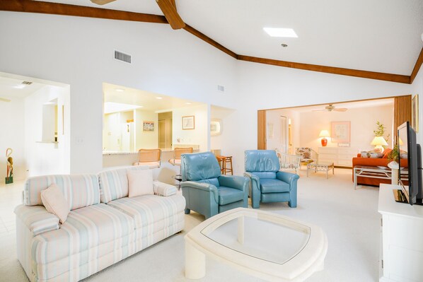 A spacious living room with a striped sofa, two blue armchairs, a glass coffee table, and a television.