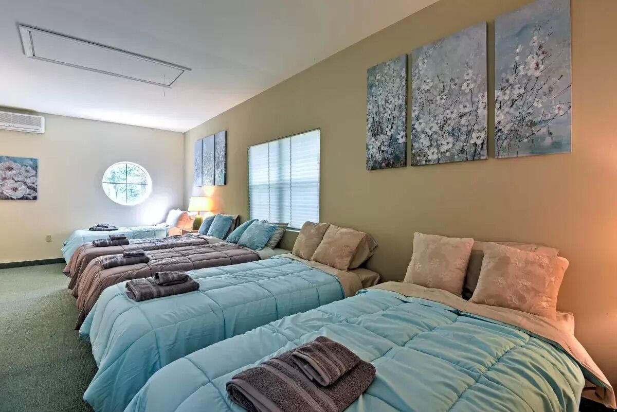 A hotel room with four beds, each with a blue quilt and a brown towel. There are three paintings on the wall, a round mirror, and a window with blinds.