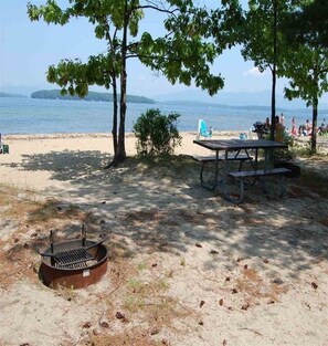 Een strand met een picknicktafel, een barbecue en een vuurkuil.