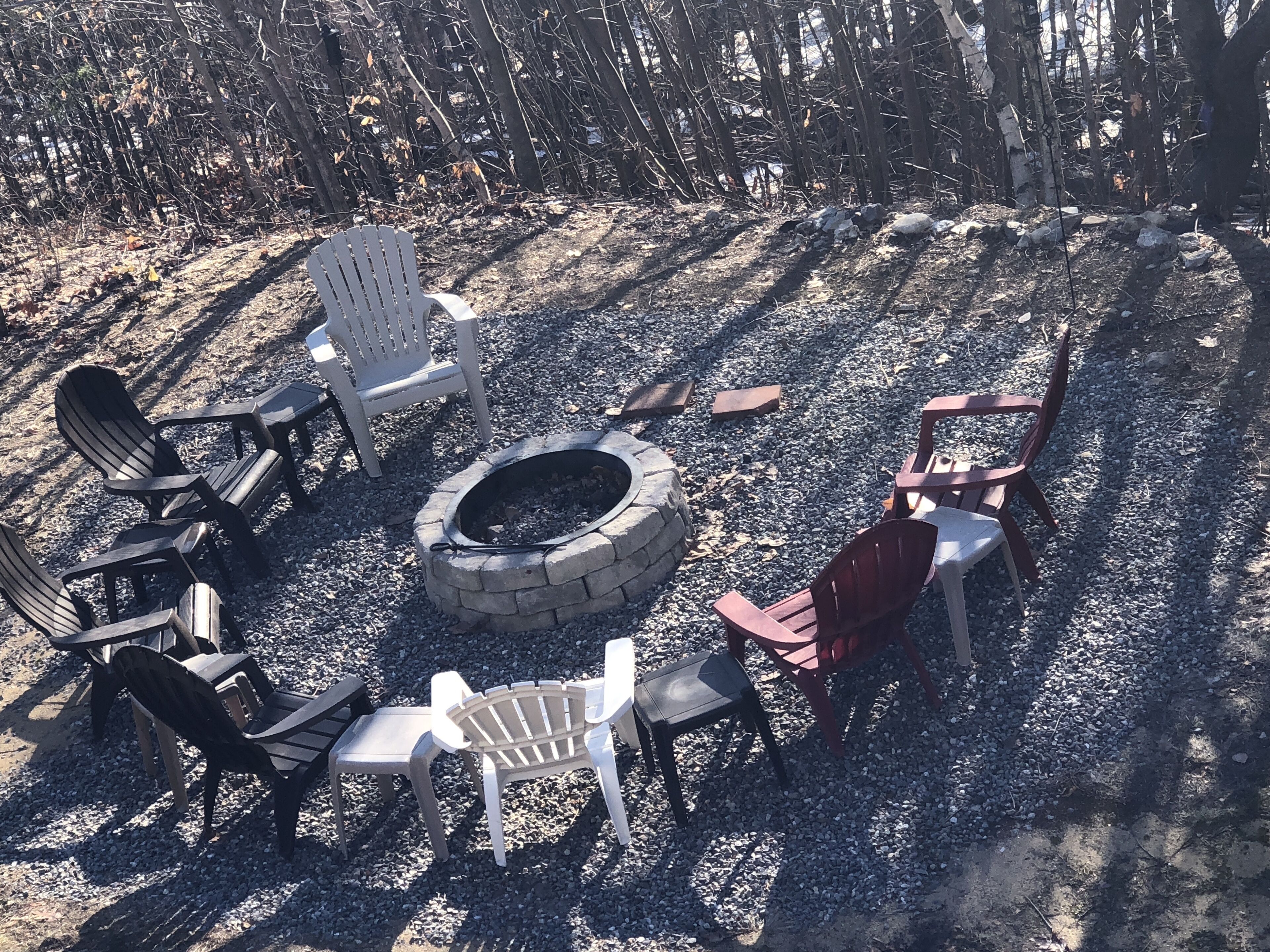 A circular stone fire pit surrounded by Adirondack chairs and a small table, set on a gravel surface with trees in the background.