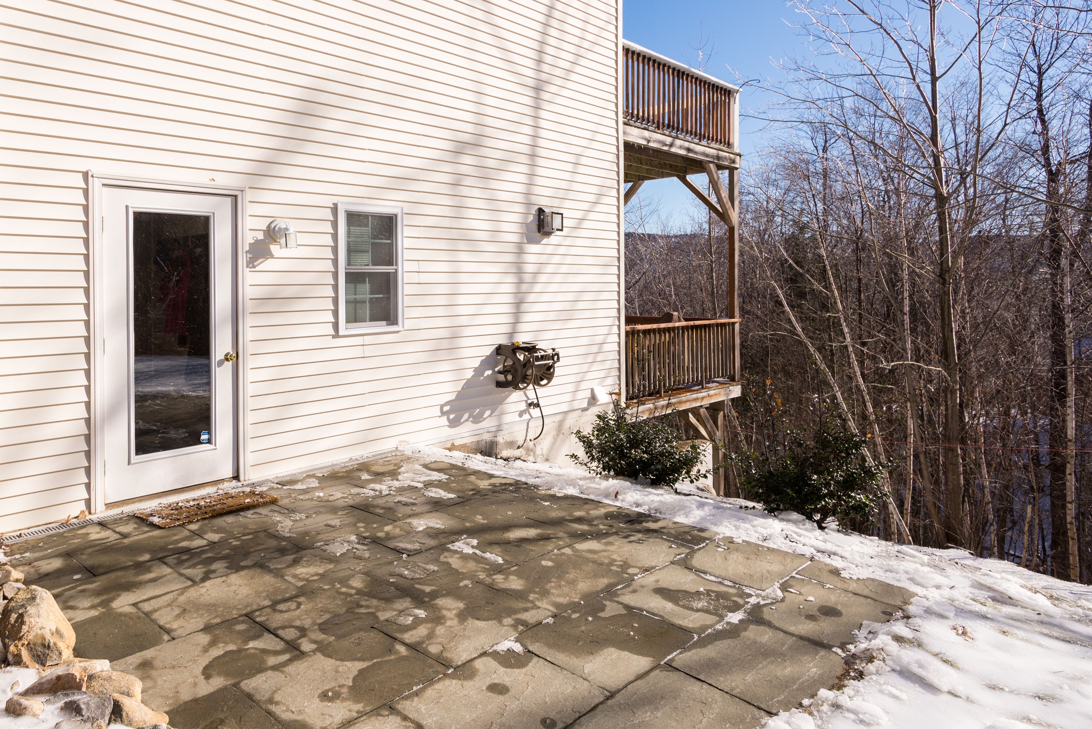 A house with a stone patio, a wooden deck, and a view of the surrounding forest.