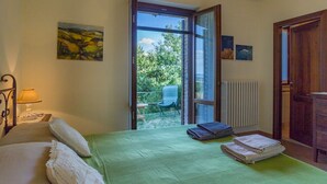 Een slaapkamer met een bed, nachtkastje, lamp, een groene plaid, een raam met open deur, een stoel erbuiten en een radiator.