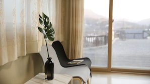Una habitación con vistas a un paisaje urbano a través de un ventanal grande, una silla negra, una mesita con un florero y una cortina con estampado floral.