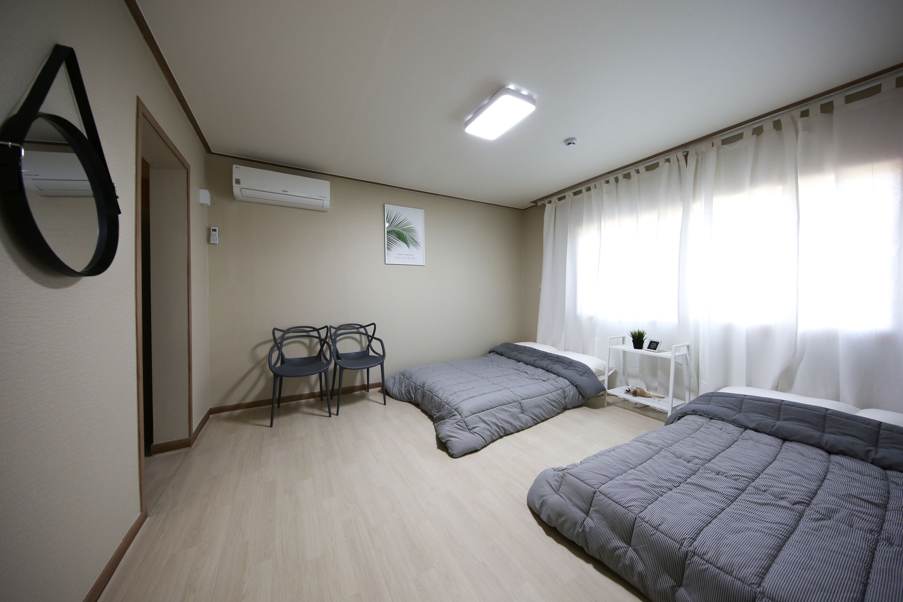 A bedroom with two beds, a desk with chairs, an air conditioner, and a window with curtains.