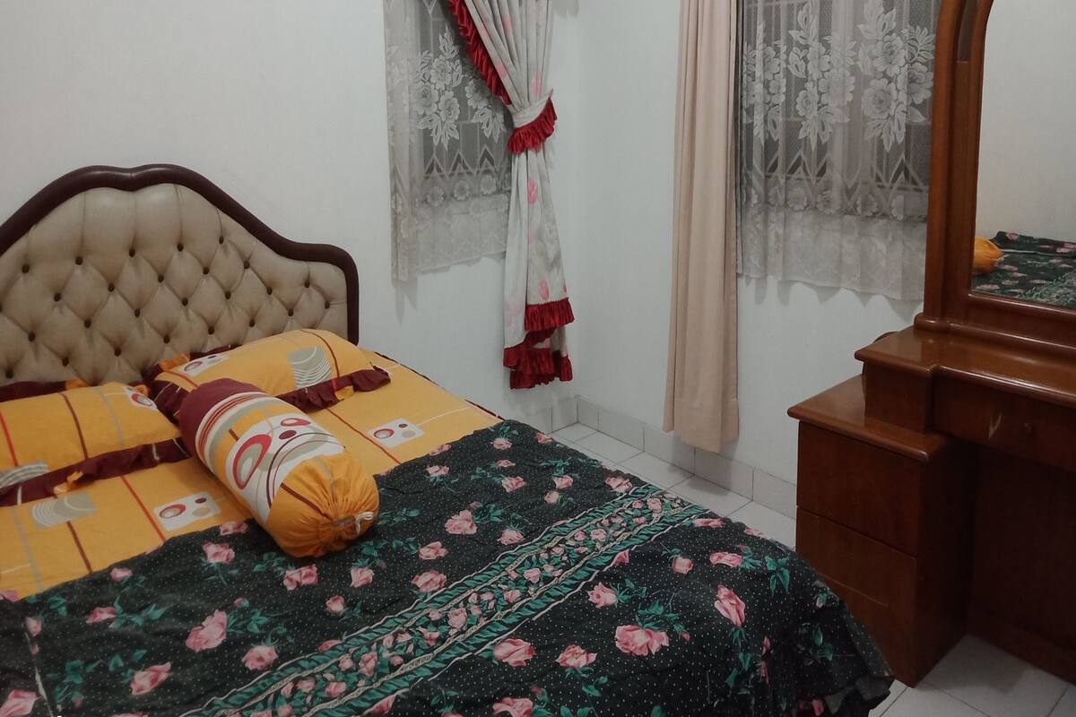 Une chambre à coucher avec une tête de lit capitonnée, un lit recouvert d’une literie à motifs floraux, une commode en bois et une fenêtre ornée de rideaux à fleurs.
