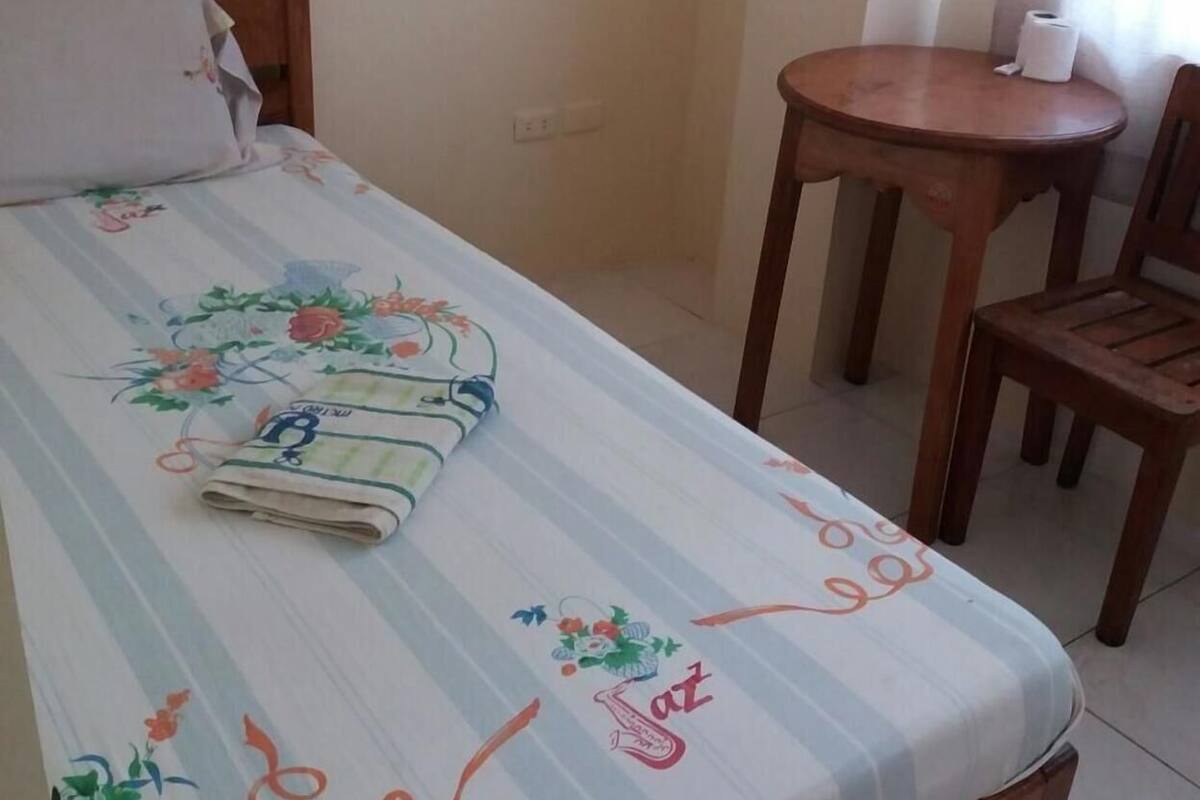 A bed with a floral bedspread, a wooden headboard, a small wooden table with a chair, and a wall socket.