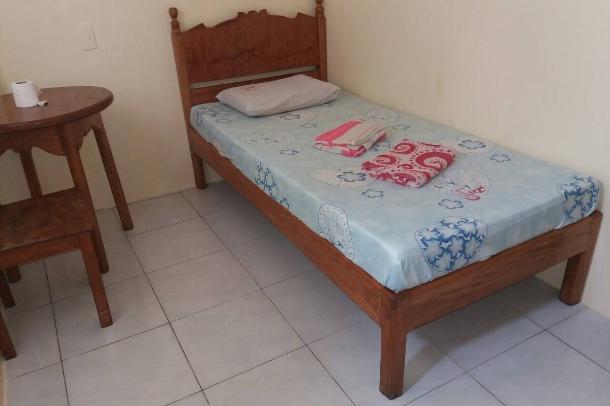 A small, neatly arranged room with a single bed, a wooden nightstand, and a chair.
