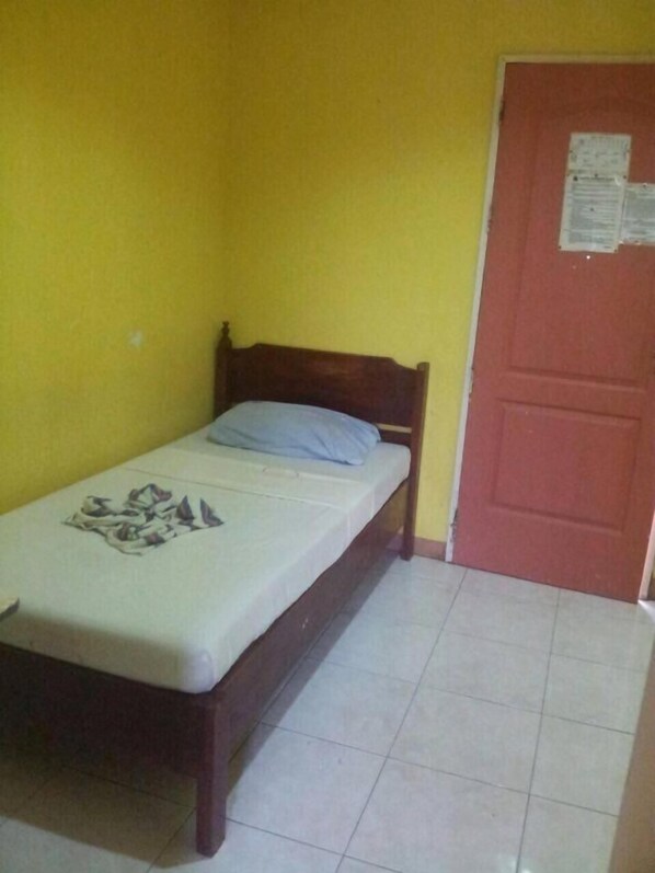 Una habitación pequeña con cama individual, cabecera de madera, colchón sencillo y una puerta roja con un aviso.