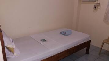 Una cama individual con colchón blanco y estructura de madera, una almohada pequeña y una manta doblada sobre la cama. Hay un equipo de aire acondicionado instalado en la pared encima de la cama.