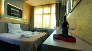 A hotel room with a bed, a bedside table with a wine bottle and glasses, a painting on the wall, and a window with curtains. - Hotel Alabin Central (Sofia)