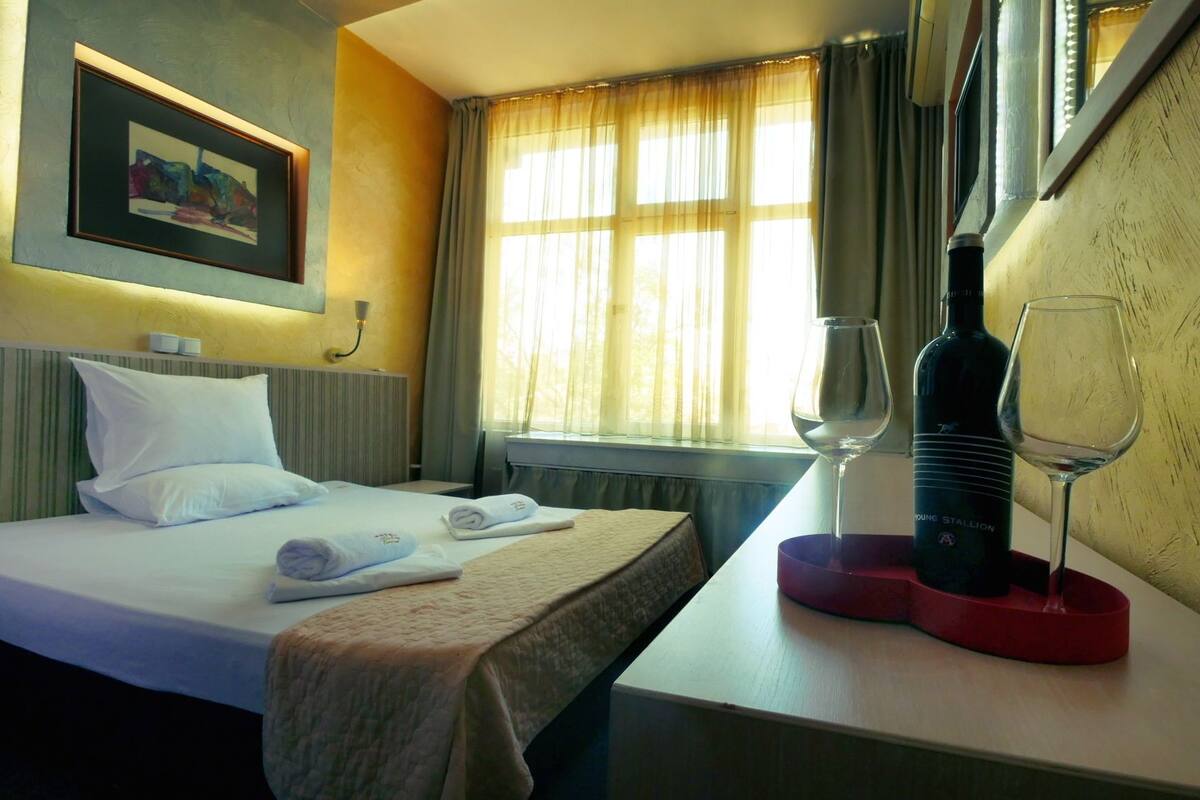 A hotel room with a bed, a bedside table with a wine bottle and glasses, a painting on the wall, and a window with curtains.