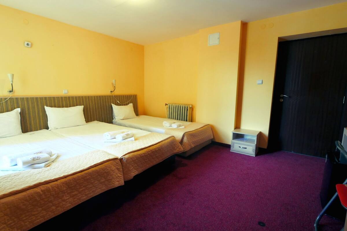 A hotel room with two beds, a small bedside table, a radiator, and a door.